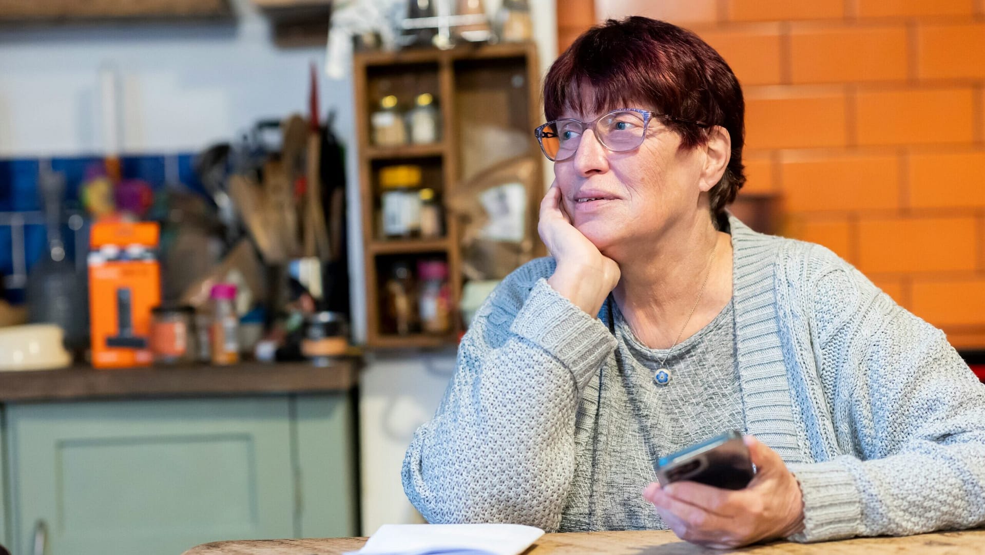 Older woman at her kitchen table holding her phone, thinking over a Medicaid planning fee quote